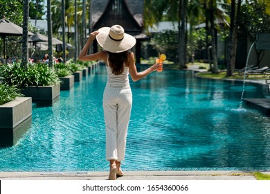 Female brunette in hat (no face), with resort style clothes white color on her slim body, enjoying holiday in tropic garden. She standing near blue swimming pool, holding glass of fresh orange drink   - Powered by Shutterstock