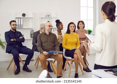 Female Business Coach In Meeting In Office Speaks In Front Of Small Group Of Business People. Group Of Multiracial Diverse Contented People Sitting On Chairs Attentively Listening To Their Leader.