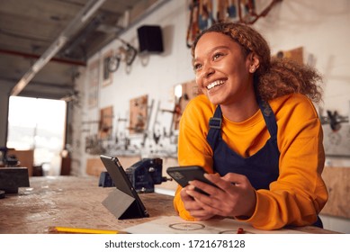 Female Business Owner In Workshop Using Digital Tablet And Holding Mobile Phone