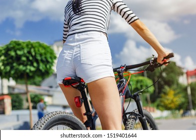 Female Buttocks In White Shorts On A Bicycle Saddle On A Bicycle Ride In The Park On A Background Of Ornamental Trees.