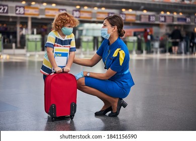 Female Cabin Crew Kneeling Before An Unaccompanied Child With One Rolling Bag While Putting Her Hand On His Shoulder