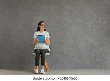 Female candidate waiting for job interview in corridor. Studio shot of unemployed young woman in glasses sitting on chair, holding CV and looking away at blank text copy space background of grey wall