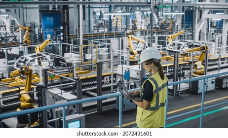 Female Car Factory Engineer In High Visibility Vest Using Laptop Computer. Automotive Industrial Manufacturing Facility Working On Vehicle Production With Robotic Technology. Automated Assembly Plant.