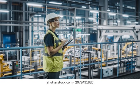 Female Car Factory Engineer in High Visibility Vest Using Laptop Computer. Automotive Industrial Manufacturing Facility Working on Vehicle Production with Robotic Arms. Automated Assembly Plant. - Shutterstock ID 2072431820