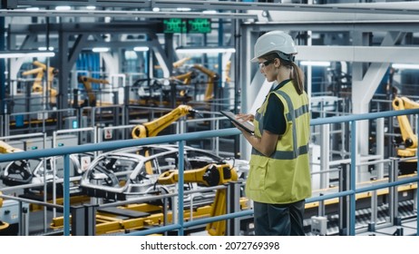 Female Car Factory Engineer In High Visibility Vest Using Tablet Computer. Automotive Industrial Manufacturing Facility Working On Vehicle Production With Robotic Technology. Automated Assembly Plant.