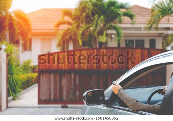 Female in car hand holding and using remote\
control to open the automatic wooden gate with modern home blurred\
background. Security and save time\
concept.