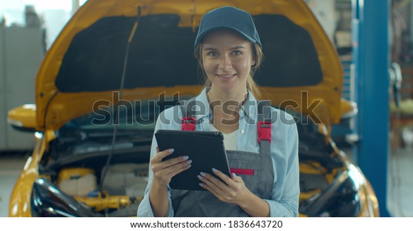 Female car mechanic using tablet device in big\
garage with cars. Beautiful woman using tablet, then smiling to\
camera in car workshop.