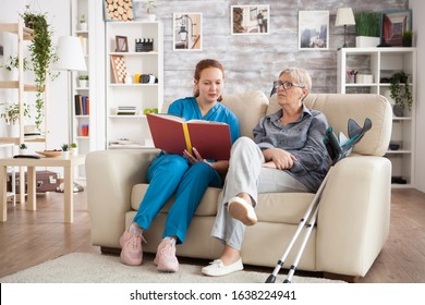 Female Care Giver Reading A Story From A Book To A Senior Woman In Nursing Home.