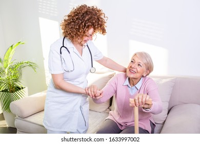 Female Caregiver Helping Senior Woman Get Up From Couch In Living Room. Smiling Nurse Assisting Senior Woman To Get Up. Caring Nurse Supporting Patient While Getting Up From Sofa