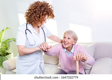 Female Caregiver Helping Senior Woman Get Up From Couch In Living Room. Smiling Nurse Assisting Senior Woman To Get Up. Caring Nurse Supporting Patient While Getting Up From Sofa