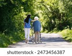Female caregiver and senior woman with walker on walk in nature. Nurse and elderly woman enjoying a warm day in nursing home, public park.
