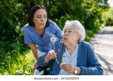 Female caregiver and senior woman in wheelchair holding dandelion, picking wild flowers. Nurse and elderly woman enjoying a warm day in nursing home, public park. - Powered by Shutterstock