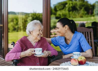 Female caregiver spending time with elderly woman, drinking coffee and talking. Nurse and senior woman in wheelchair enjoying a warm day outdoors. - Powered by Shutterstock