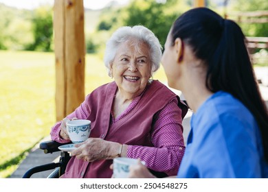 Female caregiver spending time with elderly woman, drinking coffee and talking. Nurse and senior woman in wheelchair enjoying a warm day outdoors. - Powered by Shutterstock