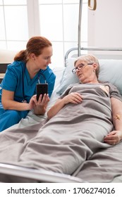 Female Caretaker In Nursing Home Talking With Old Woman Sitting In Bed And Using Mobile Phone.