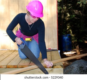 Female Carpenter Sawing Wood With Pink Hardhat, Safety Glasses And Toolbelt