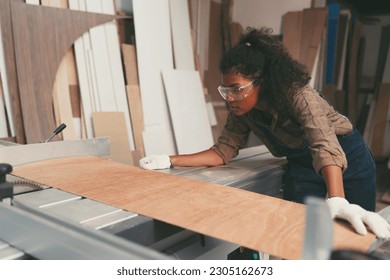 Female carpenter using saw machine for cutting sawn plywood during working in wood workshop. Female joiner wearing safety uniform and working in furniture workshop - Powered by Shutterstock