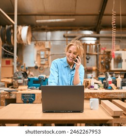 Female Carpenter Working In Woodwork Workshop Talking On Mobile Phone And Using Laptop - Powered by Shutterstock