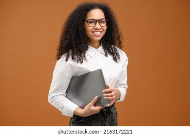 Female Carrying Laptop Computer, Smiling. Young African American Woman With Afro Hairstyle Wearing Smart Casual Wear And Stylish Eyeglasses Standing Isolated On Brown. Office Employee, Student Concept