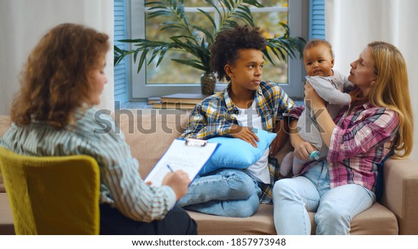 Female case worker\
visiting woman with adopted children. Back view of woman social\
carer talking to caucasian foster mother and mixed-race boy and\
baby in living room