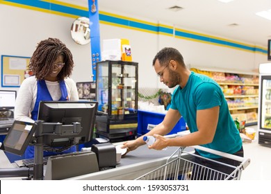 Female Cashier Reading Barcodes At Cash Register. Male Customer In Casual Putting Products From Cart To Cashier Counter. Payment In Supermarket Concept