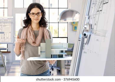 Female Casual Caucasian Architect Designer With Building Model Holding In Hand, Standing, Smiling, Looking At Camera. Bright Office.