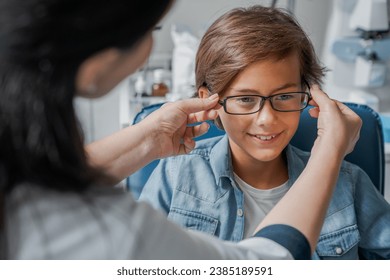 Female caucasian doctor putting glasses on little boy in clinic. Optician ophthalmologist helping assisting kid patient with selecting glasses goggles eyesight vision frame lenses - Powered by Shutterstock