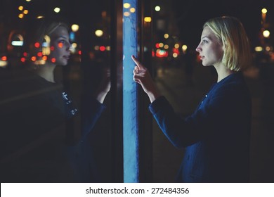 Female caucasian tourist touching information kiosk digital screen while standing outdoors at night city with beautiful lights on background, attractive young woman consults on modern big timetable