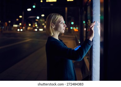Female caucasian tourist touching information kiosk digital screen while standing outdoors at night city and holding smart phone in the hand, attractive young woman consults on modern big timetable