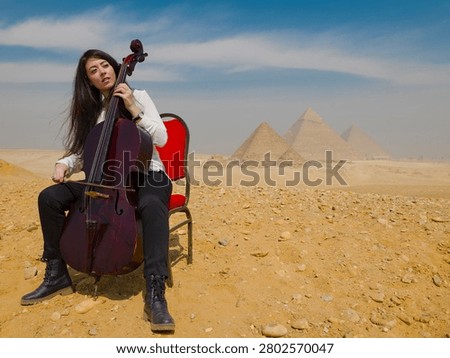 Female cellist performing cello while seated on red chair in the Giza desert before the ancient pyramids of Egypt ideal for travel music and cultural