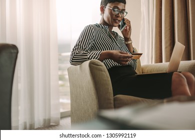 Female CEO sitting in hotel room with laptop and credit card talking over cell phone. African businesswoman doing online bill payments from hotel room.