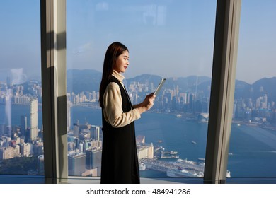 Female CEO Is Watching News In Internet Via Touch Pad, While Is Standing In Office Interior Near Window With View Of Business District. Young Chinese Woman Manager Is Using For Work Digital Tablet