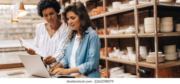 Female ceramists using a laptop while working together in their store. Female entrepreneurs managing online orders on their website. Two businesswomen running a creative small business together. - Powered by Shutterstock