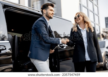 Female chauffeur greets the businessman while helping him to get out of the minivan taxi. Concept of personal driver, luxury taxi for business people