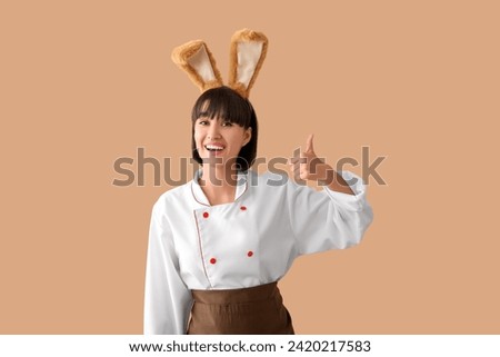 Female chef in bunny ears showing thumb-up on brown background. Easter celebration