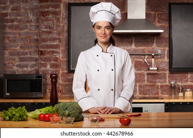 Female chef cook in a white hat in the kitchen preparing a meal from salad leaves, broccoli, tomatoes and pepper