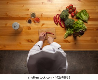 Female Chef Cutting Pepper On Wooden Cutting Board On Wooden Table With Broccoli, Salad Leaves, Tomatoes And Pepper, Top View 