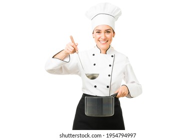 Female Chef Making Eye Contact While Holding A Ladle And A Pot With Soup. Hispanic Cooker Tasting The Cream She Just Made At A Restaurant