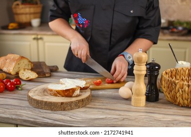 Female chef preparing breakfast in a bright country kitchen. He cuts the bread and spreads it with cheese. Kitchen table with flowers and kitchen board, bread, tomatoes, greens