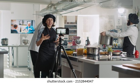 Female Chef Recording Cooking Show Video On TV Program, Filming Culinary Vlog With Camera In Professional Restaurant Kitchen. Woman In Uniform Talking About Gastronomy At Online Cuisine Class.