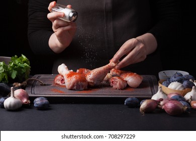 A female chef sprinkles fresh raw chicken drumsticks on a dark background with sea salt. Nearby lie the ingredients for cooking: shallots, blue plums, garlic pepper, salt and parsley. - Shutterstock ID 708297229