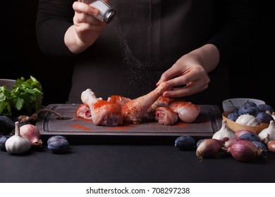 A female chef sprinkles fresh raw chicken drumsticks on a dark background with sea salt. Nearby lie the ingredients for cooking: shallots, blue plums, garlic pepper, salt and parsley. - Shutterstock ID 708297238