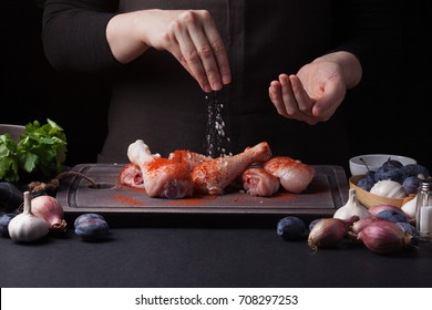 A female chef sprinkles fresh raw chicken drumsticks on a dark background with sea salt. Nearby lie the ingredients for cooking: shallots, blue plums, garlic pepper, salt and parsley. - Shutterstock ID 708297253