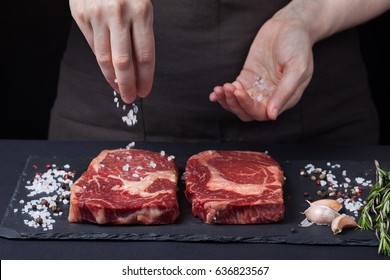 A female chef sprinkles sea salt with two fresh raw ribeye steaks from marbled beef on a dark background. Nearby is a mixture of peppers, garlic and rosemary. - Powered by Shutterstock