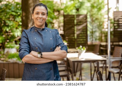 Female chef wears blue coat, happy smiling poses with arms crossed outdoors on the terrace of a restaurant. Restaurant worker, culinary gourmet, pastry chef. - Powered by Shutterstock