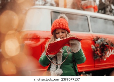 Female child celebrating Christmas and New Year winter holidays season outdoor. Little girl joyful spending time on open air holding letter to Santa in red envelope and waiting for miracle - Powered by Shutterstock