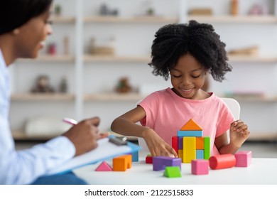 Female child development specialist observing cheerful child preschooler, happy little african american girl sitting at table and making pyramids from colorful wood blocks. Child mental health concept - Shutterstock ID 2122515833