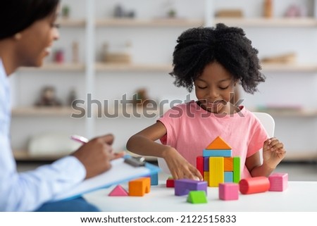 Female child development specialist observing cheerful child preschooler, happy little african american girl sitting at table and making pyramids from colorful wood blocks. Child mental health concept