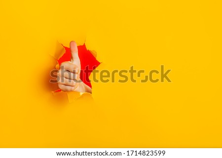 Female child hand gesturing the ok sign, like gesture through torn yellow paper wall, keeps thumb up. Approval sign, recommends something. Copy space fo text information. Best choice concept.