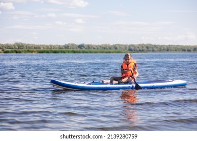 Female child sup boarding on her own on calm lake with oar in hands looking at water ripples in vest life jacket. Active holidays. Inculcation of love for sports from childhood. - Shutterstock ID 2139280759
